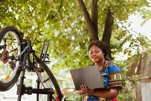 gezond toegewijd Afrikaanse Amerikaans vrouw zoeken Aan laptop naar fix gebroken modern fiets. actief vrouw fietser waarborgt fiets componenten zijn beveiligen voor zomer buitenshuis vrije tijd wielersport. foto