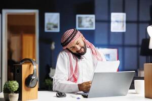 Arabisch mannetje freelancer ijverig multitasking met laptop en mobiele telefoon. maken belt, controle aantekeningen en dirigeren online Onderzoek, moslim Mens omarmt modern technologie en effectief communicatie. foto