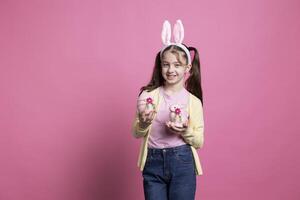 weinig meisje met pluizig konijn oren houdt roze Pasen regelingen in voorkant van camera, tonen haar versierd ei en gevuld konijn speelgoed. jong kind glimlachen in studio en vieren april evenement. foto