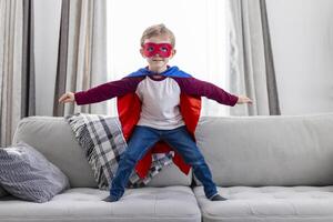 jongen in superheld kostuum met kaap en masker staand Aan sofa foto