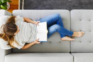 top visie van jong brunette vrouw ontspannende Aan sofa en lezing roman boek Bij huis foto