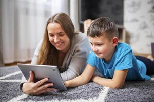 gelukkig liefhebbend familie. jong moeder en haar zoon gebruik makend van tablet aan het liegen Aan tapijt foto