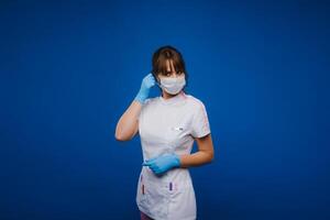 een dokter meisje staat in een medisch masker Aan een geïsoleerd blauw achtergrond foto