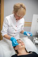 dokter schoonheidsspecialist of dermatoloog maken gezicht masker in schoonheidsspecialiste salon. professioneel schoonheidsspecialist toepassen gezicht masker Aan Kaukasisch vrouw gezicht aan het liegen Aan bed in badjas. foto