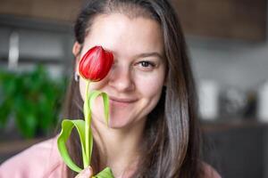 portret van glimlachen jong vrouw met rood tulp Bij huis. focus Aan tulp. Gefeliciteerd en een geschenk Aan Internationale vrouwen dag of moeder dag foto