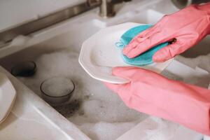 huisvrouw meisje in roze handschoenen wast gerechten door hand- in de wastafel met wasmiddel. de meisje reinigt de huis en wast gerechten in handschoenen Bij huis. foto