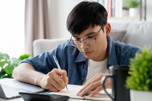 de Mens in gewoontjes kleren werken met een laptop, computer, slim telefoon, rekenmachine zittend Aan de sofa in de leven kamer Bij huis, werken van huis concept. foto