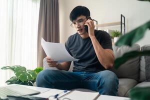 de Mens in gewoontjes kleren werken met een laptop, computer, slim telefoon, rekenmachine zittend Aan de sofa in de leven kamer Bij huis, werken van huis concept. foto