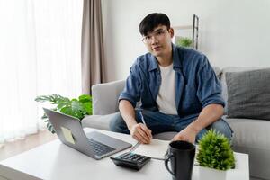 de Mens in gewoontjes kleren werken met een laptop, computer, slim telefoon, rekenmachine zittend Aan de sofa in de leven kamer Bij huis, werken van huis concept. foto