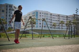 jong atletisch Mens het uitvoeren van een cardio training, jumping met een overslaan touw Aan de buitenshuis stad sportveld foto
