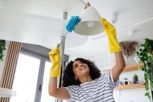 jong huishoudster in schort Afstoffen de kroonluchter door stofdoek voorzichtig schoonmaak in leven kamer Bij huis. jong vrouw in rubber handschoenen aan het doen huiswerk. foto