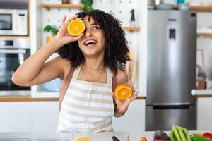 foto van schattig Afrikaanse Amerikaans vrouw glimlachen en Holding twee oranje onderdelen terwijl Koken groente salade in keuken interieur Bij huis