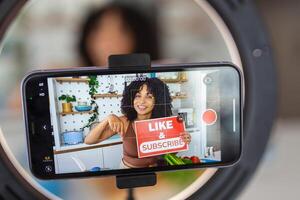 een Afro-Caribisch vrouw tonen een creatief recept met gezond vers ingrediënten van haar keuken Bij huis, gebruik makend van haar slim telefoon naar film de kookkunst demonstratie foto