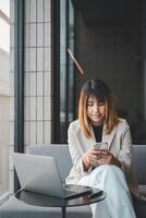 een gefocust ondernemer in een chique blazer concentraten Aan haar smartphone, beheren haar bedrijf zaken met een laptop dichtbij, in een modern werkruimte instelling. foto