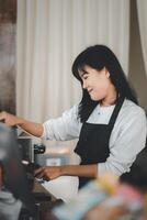 een glimlachen barista in een zwart schort regelt cups Bij een koffie winkel, haar aangenaam houding toevoegen warmte naar de sfeer. foto