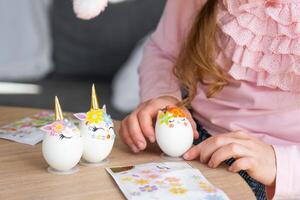 een schattig meisje met roze konijn oren maakt een Pasen ambacht - siert een ei in de het formulier van een eenhoorn met strass steentjes, Hoorn, bloemen in de interieur van een huis met planten. foto
