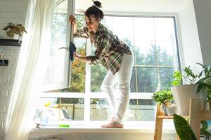 vrouw handmatig wast de venster van de huis met een vod met verstuiven schoonmaakster en dweilen binnen de interieur met wit gordijnen. herstellen bestellen en netheid in de lente, schoonmaak onderhoud foto