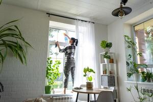 vrouw handmatig wast de venster van de huis met een vod met verstuiven schoonmaakster en dweilen binnen de interieur met wit gordijnen. herstellen bestellen en netheid in de lente, schoonmaak onderhoud foto