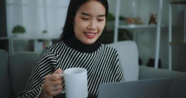 portret van gelukkig jong Aziatisch vrouw zitten Aan sofa drinken koffie of thee terwijl werken creatief baan van huis Aan laptop met online netwerk, nemen pauze, glimlachend foto