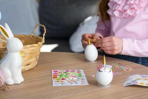 een schattig meisje met roze konijn oren maakt een Pasen ambacht - siert een ei in de het formulier van een eenhoorn met strass steentjes, Hoorn, bloemen in de interieur van een huis met planten. foto