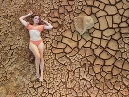 antenne portret van mooi jong dame in bikini aan het liegen Aan droog gebarsten rood bodem. dor, gebarsten rood bodem droogte en dor klimaat conditie. klimaat en weer Wijzigen. foto