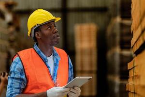 Afrikaanse arbeider timmerman vervelend veiligheid uniform en moeilijk hoed werken en controle de kwaliteit van houten producten Bij werkplaats productie. Mens en vrouw arbeiders hout in donker magazijn industrie. foto
