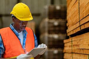 Afrikaanse arbeider timmerman vervelend veiligheid uniform en moeilijk hoed werken en controle de kwaliteit van houten producten Bij werkplaats productie. Mens en vrouw arbeiders hout in donker magazijn industrie. foto