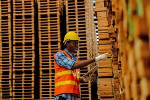 Afrikaanse arbeider timmerman vervelend veiligheid uniform en moeilijk hoed werken en controle de kwaliteit van houten producten Bij werkplaats productie. Mens en vrouw arbeiders hout in donker magazijn industrie. foto