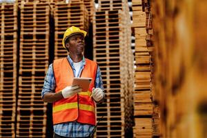 Afrikaanse arbeider timmerman vervelend veiligheid uniform en moeilijk hoed werken en controle de kwaliteit van houten producten Bij werkplaats productie. Mens en vrouw arbeiders hout in donker magazijn industrie. foto