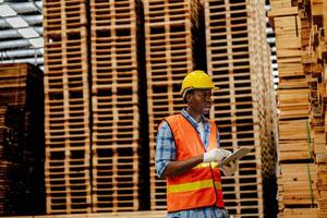 Afrikaanse arbeider timmerman vervelend veiligheid uniform en moeilijk hoed werken en controle de kwaliteit van houten producten Bij werkplaats productie. Mens en vrouw arbeiders hout in donker magazijn industrie. foto