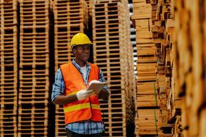 Afrikaanse arbeider timmerman vervelend veiligheid uniform en moeilijk hoed werken en controle de kwaliteit van houten producten Bij werkplaats productie. Mens en vrouw arbeiders hout in donker magazijn industrie. foto