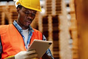 Afrikaanse arbeider timmerman vervelend veiligheid uniform en moeilijk hoed werken en controle de kwaliteit van houten producten Bij werkplaats productie. Mens en vrouw arbeiders hout in donker magazijn industrie. foto