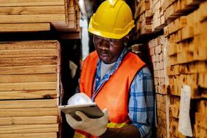 Afrikaanse arbeider timmerman vervelend veiligheid uniform en moeilijk hoed werken en controle de kwaliteit van houten producten Bij werkplaats productie. Mens en vrouw arbeiders hout in donker magazijn industrie. foto