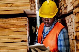 Afrikaanse arbeider timmerman vervelend veiligheid uniform en moeilijk hoed werken en controle de kwaliteit van houten producten Bij werkplaats productie. Mens en vrouw arbeiders hout in donker magazijn industrie. foto