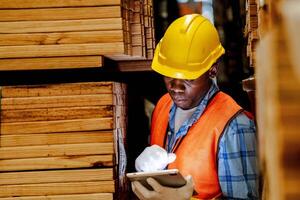 Afrikaanse arbeider timmerman vervelend veiligheid uniform en moeilijk hoed werken en controle de kwaliteit van houten producten Bij werkplaats productie. Mens en vrouw arbeiders hout in donker magazijn industrie. foto