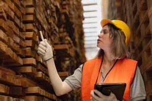 vrouw arbeider timmerman vervelend veiligheid uniform en moeilijk hoed werken en controle de kwaliteit van houten producten Bij werkplaats productie. Mens en vrouw arbeiders hout in donker magazijn industrie. foto