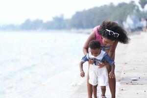 gemengd ras Afrikaanse en Aziatisch moeder en jongen is spelen Bij de buitenshuis Oppervlakte. glimlachen gelukkig familie hebben pret rennen Aan de strand. portret van mam en kind levensstijl met een uniek kapsel. foto