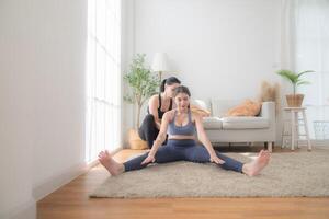 twee Dames zelfverzekerd opleiding yoga. atletisch Dames in sportkleding aan het doen geschiktheid uitrekken opdrachten Bij huis in de leven kamer. sport en recreatie concept. yoga leraar is helpen jong vrouw. foto