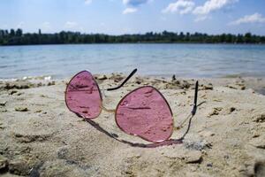 roze bril Aan de zand in de buurt blauw water van meer. foto