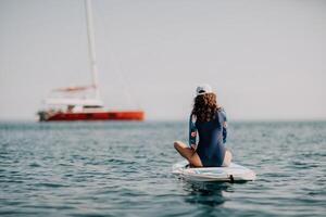 vrouw sup yoga. midden- leeftijd sportief vrouw beoefenen yoga pilates Aan peddelen sup surfplank. vrouw uitrekken aan het doen training Aan zee water. modern individu vrouw hipster buitenshuis zomer sport werkzaamheid. foto