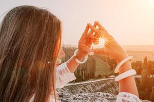 gelukkig vrouw in wit boho jurk maken hart teken met handen Aan zonsondergang in bergen. romantisch vrouw met lang haar- staand met haar terug Aan de zonsondergang in natuur in zomer met Open handen. foto