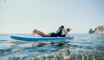 zee vrouw sup. silhouet van gelukkig positief jong vrouw met haar hond, surfing Aan sup bord door kalmte water oppervlak. idyllisch zonsondergang. actief levensstijl Bij zee of rivier. zomer vakantie met huisdieren. foto