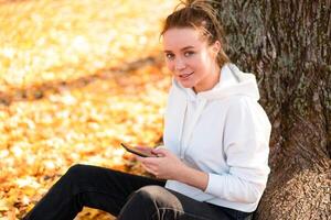 vrouw in een wit trui met een kap zit Aan grond in de park in de buurt de boom en Holding een mobiele telefoon in haar handen foto