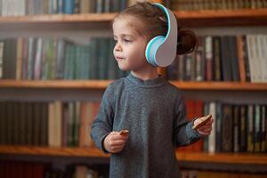 schattig Kaukasisch weinig meisje in groot wit hoofdtelefoons luistert muziek- in de interieur tegen de achtergrond van boekenplanken. foto