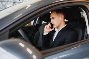 jong zakenman zit in luxe auto en praat Aan telefoon. hij looks Rechtdoor naar voren. vent schijven auto. hij houdt een hand- Aan stuurinrichting wiel. het is zonnig buiten. foto