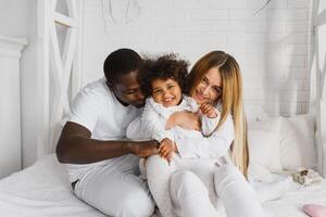 portret van gelukkig multiraciaal jong familie aan het liegen Aan knus wit bed Bij huis, glimlachen Internationale mam en vader ontspannende met weinig biraciaal meisje kind poseren voor afbeelding in slaapkamer foto