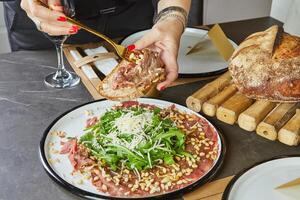 een elegant gastvrouw in een modern keuken elegant plaatsen een heerlijk schotel van carpaccio Aan brood Aan een schoon marmeren aanrecht, demonstreren de kunst van presentatie en culinaire dapperheid foto