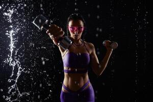 mooi jong meisje in Purper sportkleding poses met halters in aqua studio. druppels van water verspreiding over haar geschiktheid lichaam. de perfect figuur Aan de achtergrond van water spatten foto