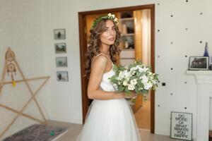 de bruid in een mooi bruiloft jurk met een lang gekruld haar- staat in de slaapkamer en looks Bij de camera. bruid binnenshuis met wit bloemen boeket en inschrijving krans kapsel foto