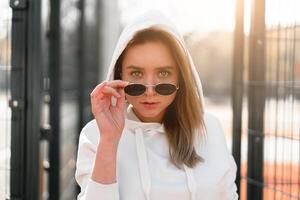 buitenshuis dichtbij omhoog portret van jong mooi vrouw met lang haar- in zonnebril, gekleed in een wit trui, in de buurt de sportveld foto